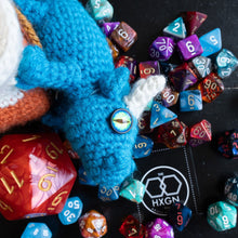 Load image into Gallery viewer, A blue knitted dragon toy with a white horn and a compass button for an eye, surrounded by colorful polyhedral gaming dice on a dark tabletop. A black square card with the white logo 'The HXGN' is visible near the bottom right.