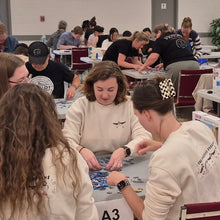 Load image into Gallery viewer, Group of people sitting at tables in a large room, solving puzzles.