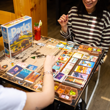 Load image into Gallery viewer, Two people playing a board game with a 'Parks' box on a wooden table.