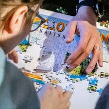 Load image into Gallery viewer, Two people working on a puzzle together, with one person's hand helping another piece fit.
