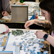 Load image into Gallery viewer, People working on a puzzle at a table with various items in the background