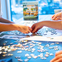 Load image into Gallery viewer, Two people working on a puzzle together with scattered pieces on a table.