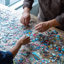 Load image into Gallery viewer, Two people working on a puzzle with colorful pieces on a wooden table.