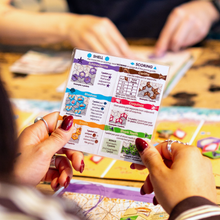 Load image into Gallery viewer, Person holding a card with game rules and scoring system in a board game setting.