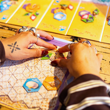 Load image into Gallery viewer, Close-up of hands playing a board game with colorful cards and hexagonal game pieces.