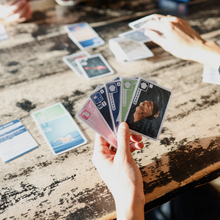 Load image into Gallery viewer, Hands holding playing cards on a wooden table with other cards displayed on a table