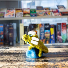 Load image into Gallery viewer, A handcrafted crochet dragon dice bag, with a dragon design clutching a bottle, displayed on a wooden surface with a shelf of books in the background.
