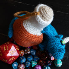 Load image into Gallery viewer, A crocheted potion bottle toy with a blue body, orange neck, and cream-colored stopper, styled with a variety of colorful polyhedral gaming dice on a dark wooden surface. A black card with the white logo 'HXGN' is visible at the bottom.
