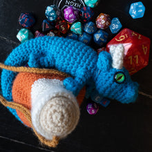 Load image into Gallery viewer, A crocheted blue dragon figurine with orange, white, and tan details, posed on a dark wooden surface and surrounded by colorful polyhedral dice.
