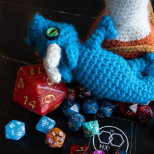 Load image into Gallery viewer, A blue crocheted dragon toy with white horns and a green eye lies on a dark tabletop surrounded by colorful polyhedral gaming dice and a black dice-rolling mat.
