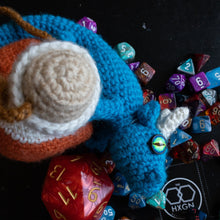 Load image into Gallery viewer, A crocheted blue dragon toy with a white horn and a small green eye, styled on a black background with colorful polyhedral dice and a black square card printed with the brand logo 'HXGN'.
