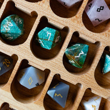 Load image into Gallery viewer, A close-up of a wooden compartment organizer holding multiple polyhedral dice. The dice include green marbled d20s, pale pink translucent d12s, and gray/white dice with gold and black markings, arranged in a light natural wood tray.
