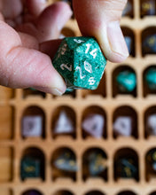 Load image into Gallery viewer, A close-up of a hand holding a green polyhedral die with white numbering, with a blurred wooden tray of other dice in the background.
