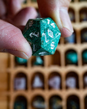 Load image into Gallery viewer, A close-up of a hand holding a green d20 polyhedral die with white numbering, with a blurred background of a wooden tray holding more dice.
