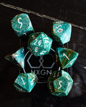 Load image into Gallery viewer, A set of seven green and black mottled polyhedral dice with white numbering, arranged on a dark marble surface around a branded black mat.
