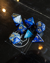 Load image into Gallery viewer, A set of blue and gold polyhedral tabletop gaming dice arranged on a dark tabletop, with one die resting on a branded game cafe mat.
