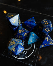 Load image into Gallery viewer, A set of blue polyhedral gaming dice with gold numbering, including a d20 and several other standard RPG dice, arranged on a dark reflective surface with a visible BN Game Cafe logo underneath.
