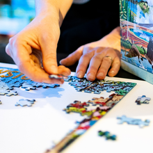 Load image into Gallery viewer, Person assembling a puzzle with a scenic box in the background

