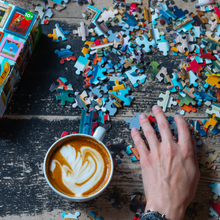 Load image into Gallery viewer, Puzzle pieces scattered on a wooden table with a hand reaching for a piece, next to a cup of coffee.
