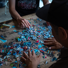 Load image into Gallery viewer, Two people working on a puzzle with colorful pieces on a wooden table.
