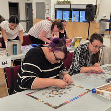 Load image into Gallery viewer, People working on puzzles at tables in a room with chairs and tables.
