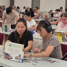Load image into Gallery viewer, Group of people sitting at tables in a large room, solving puzzles.

