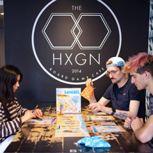 Load image into Gallery viewer, People playing board games at The HXGN Board Game Cafe with a game box titled 'Sanibel' in the foreground.
