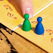 Load image into Gallery viewer, Two game pieces, one green and one blue, on a yellow game board with footprints.
