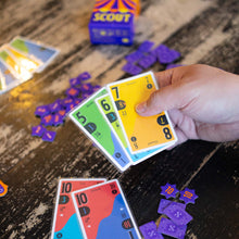 Load image into Gallery viewer, Hand holding colorful card game cards on a wooden surface with scattered game pieces.

