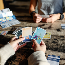 Load image into Gallery viewer, Two people playing a card game at a wooden table with various cards scattered around.
