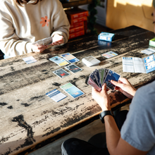 Load image into Gallery viewer, Two people playing a card game at a wooden table.
