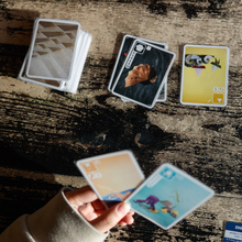 Load image into Gallery viewer, Hand holding playing cards on a wooden surface with other cards displayed on a table
