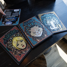 Load image into Gallery viewer, Three illustrated boxed cards or miniatures labeled 'Astrea Adventurer's Set' are arranged on a dark wooden table, with one box open to show the contents. A hand is partially visible at the left edge.

