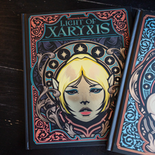 Load image into Gallery viewer, A hardcover book titled 'Light of Xaryxis' by Brandon Sanderson, featuring a highly detailed fantasy illustration of a blonde female elf on the cover, displayed on a dark wooden surface next to another book.

